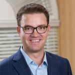 Bryan Sina photo Photo of a smiling man wearing a suit and glasses in front of a window and blinds.