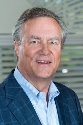 Photograph of a smiling man with in front of a window, wearing a plaid suit.