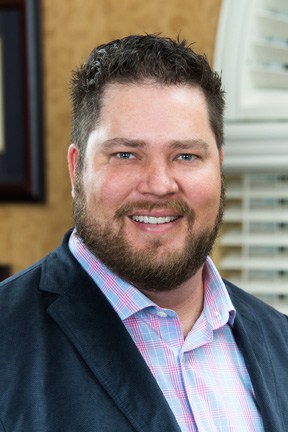 Photograph of a smiling man with a mustache and beard in front of a window, wearing a suit.