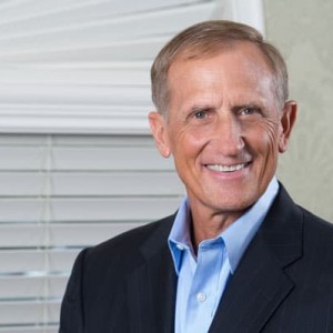 A photograph of a man wearing a suite, next to a window with blinds.