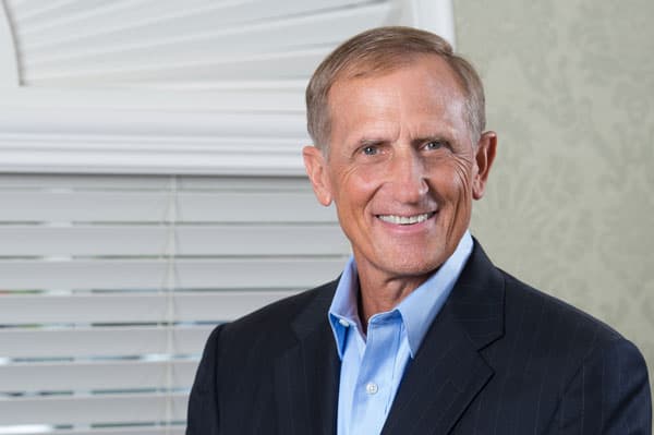 A photograph of a man wearing a suite, next to a window with blinds.